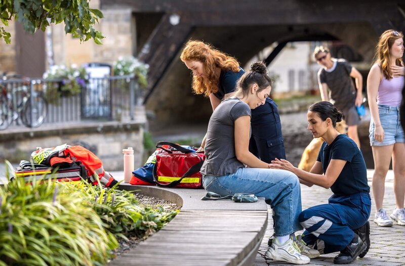 Dr. Leyla Sherbaz (Sanam Afrashteh, r.) und Tamar Hummel (Linda Kummer, l.) leisten erste Hilfe bei Gina Hachmann (Lucia E. Lampolia, M.), die sich bei einem Sturz eine Verletzung an der Hand zugezogen hat (mit Komparsen, h.). – Bild: ARD/​Michael Kremer
