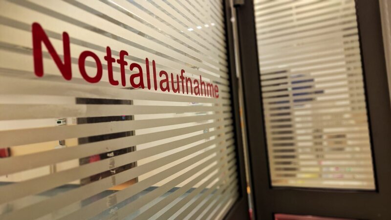 Ep2 – a glass window with the German word for ER (Notfallaufnahme) on it and stripes to provide some kind of privacy. – Bild: Warner Bros. Discovery, Inc.