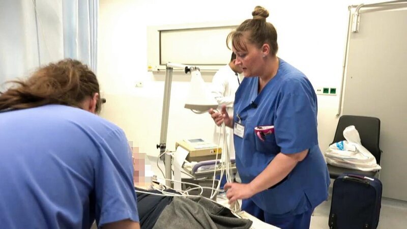 Nurse Karoline is holding some cables in her hand while apparently talking to a blurred patient. In the back we see a young doctor in a white coat, to the right a carry on bagand a chair. A male long haired nurse is seen on the left front. – Bild: Warner Bros. Discovery, Inc. or its subsidiaries and affiliates
