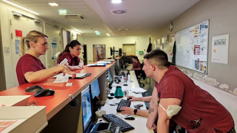 Some members of staff are talking and listening to each other at the orange counter. Y female nurse wearing glasses and a female physician are leaning on, a young male nurse with a mask hanging on his arm is listening. – Bild: DMAX