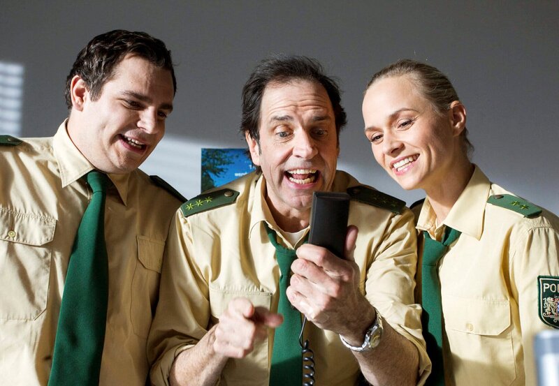 Martin Riedl (Paul Sedlmeir, l.), Johannes Staller (Helmfried von Lüttichau, M.) und Sonja Wirth (Anett Fleischer, r.) amüsieren sich mit einem Handy. – Bild: ARD/​TMG/​Chris Hirschhäuser