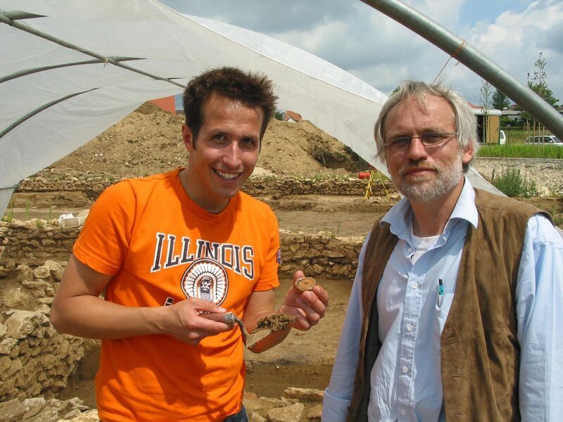 Willi Weitzel präsentiert mit dem Grabungsleiter Dr. Claus-Michael Hüssen ein Fundstück. Heute ist er dabei, wie Archäologen ein römisches Hofgut ausgraben. – Bild: BR/​megaherz gmbh