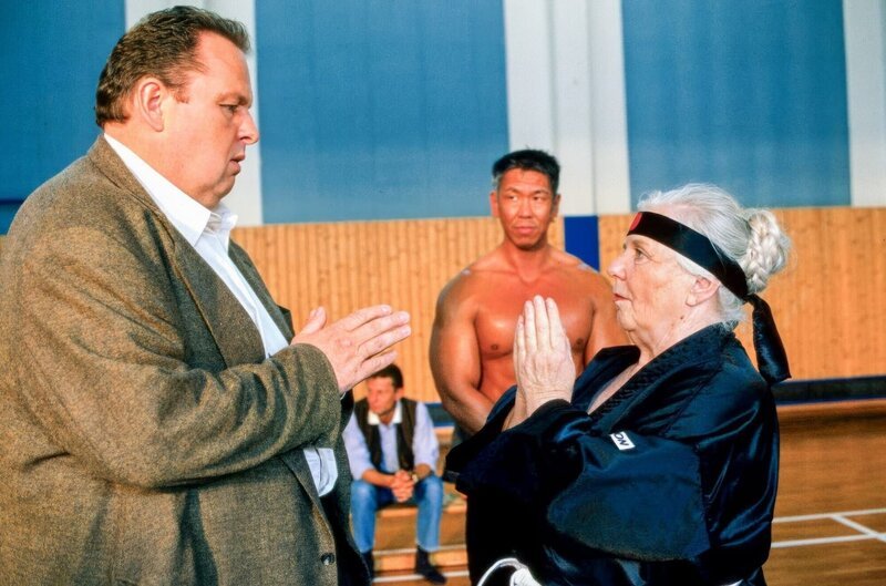 Mutter Resi (Ruth Drexel, r.) hat sich zu einem Selbstverteidigungskurs bei Bruce Adelholzer (Jocky Wong, M.) angemeldet. Stolz zeigt sie Benno (Ottfried Fischer, l.), wie man einen Kampf beginnt. – Bild: ORF/​ZDF