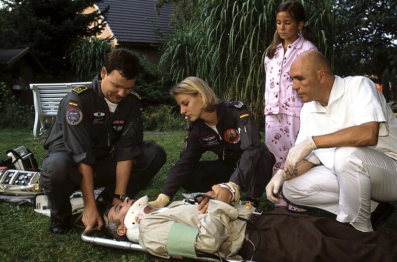 Pitter Schrankel (Dietrich Adam, 2.v.l.) ist von der Leiter gefallen, als er versuchte, die Katze seiner Tochter vom Baum zu holen. Thomas Asmus (Ulrich Bähnk, l.) und Sabine Petersen (Marlene Marlow, M.) versorgen den Verletzten, der auf einer Trage liegt. Seine Tochter (Julina Senol, 2.v.r.) steht besorgt daneben. – Bild: ZDF