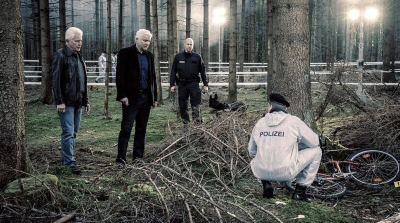 Die Kriminalhauptkommissare Ivo Batic (Miroslav Nemec) und Franz Leitmayr (Udo Wachtveitl) mit Kollegen (Komparsen) betrachten den Fundort von Emils Fahrrad im Wald, in der Nähe eines Parkplatzes. – Bild: BR/​Bavaria Fiction GmbH