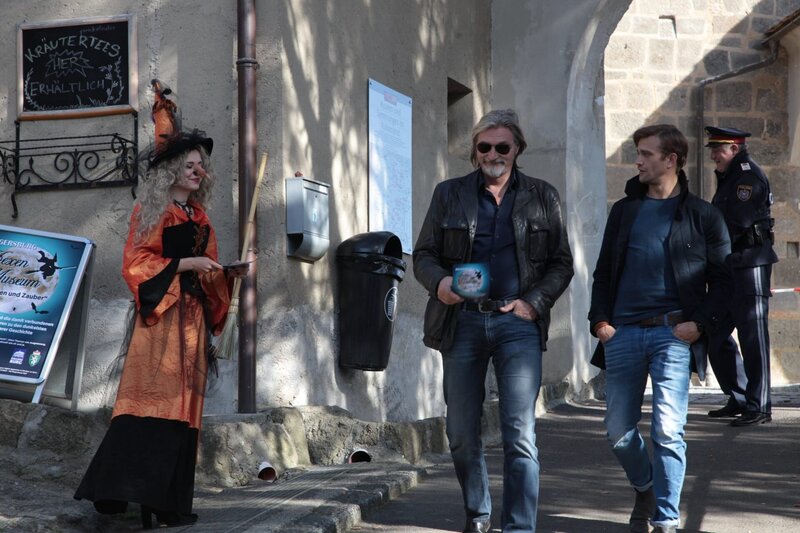 Major Carl Ribarski (Stefan Jürgens, M.) und Bezirksinspektor Simon Steininger (Michael Steinocher, r.) bekommen eine Broschüre über das Hexenmuseum in Riegersburg gereicht. (v.l.n.r.: Komparsin, Major Carl Ribarski (Stefan Jürgens), Bezirksinspektor Simon Steininger (Michael Steinocher), im Hintergrund: Leopold Moser (Karl Fischer)). – Bild: ORF/​Satel Film/​Stefan Haring
