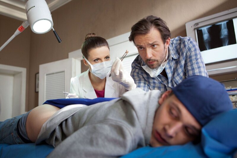 Eva (Julia Hartmann) und Adam (Lucas Gregorowicz) operieren Ronny (Rick Okon, vorne) eine Kugel aus dem Allerwertesten. – Bild: RTL