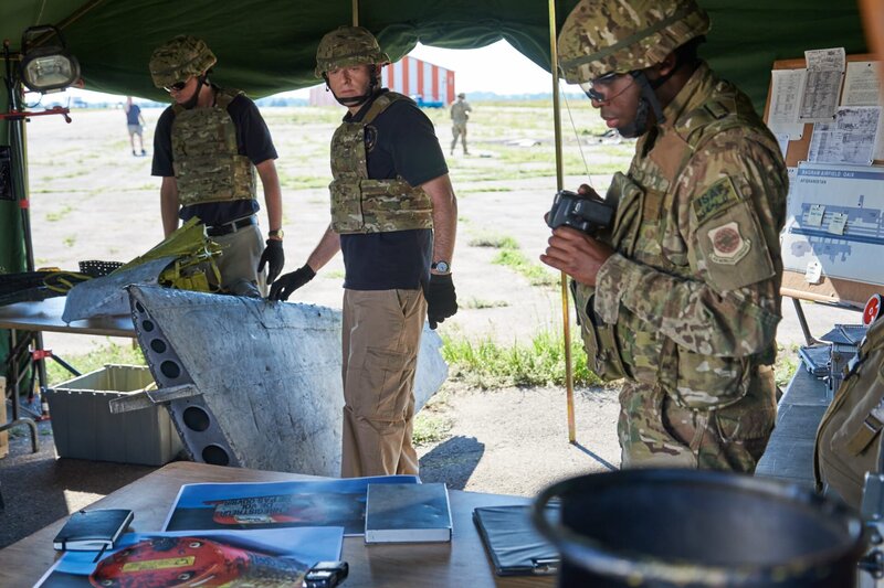 ONTARIO – RE-ENACTMENT – Investigator (played by Dmitry Chepovetsky) along with his team and military personnel (played by background performers) study pieces of wreckage in the crash site tent. – Bild: Cineflix/​Steven Lungley