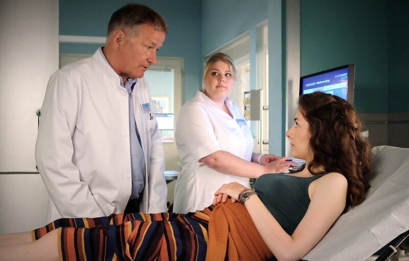 Die engagierte Anke Baumeister (Amy Benkenstein, rechts) organisiert gerade eine Spendenveranstaltung für ein Tierheim und hat nicht wirklich Zeit, sich um sich selbst zu kümmern. Doch Dr. Heilmann (Thomas Rühmann, links) muss die Lähmungserscheinungen in Ankes Beinen, die für ihren Zusammenbruch verantwortlich sind, abklären. Und dafür muss Schwester Miriam (Christina Petersen) wenigstens Blut abnehmen. – Bild: Sebastian Kiss