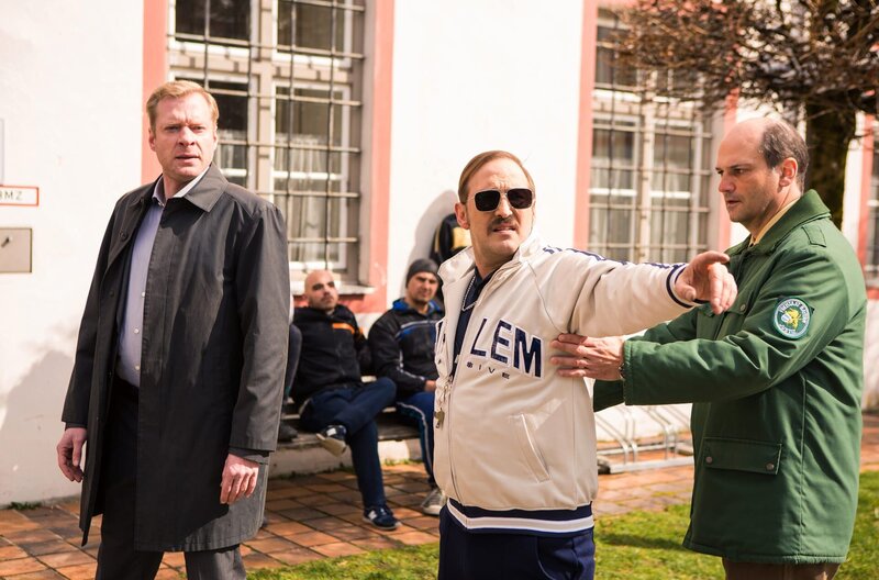 Der „geflüchtete“ Fußballtrainer (Peter Kollmann, M.) ist außer sich – keiner glaubt ihm, dass Riedl ihn angestiftet hat (Stefan Kaminsky als Anstaltsleiter, l. und Komparse, r.). – Bild: ARD/​Kai Neunert