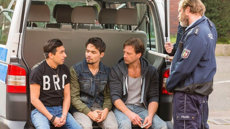 Stefan Vollmer (Hendrik Duryn, 3.v.l.) nimmt seine Schüler Mehmet (Zejhun Demirov, l.) und Jamil (Aram Arami) vor der Polizei in Schutz. – Bild: TVNOW /​ Frank Dicks