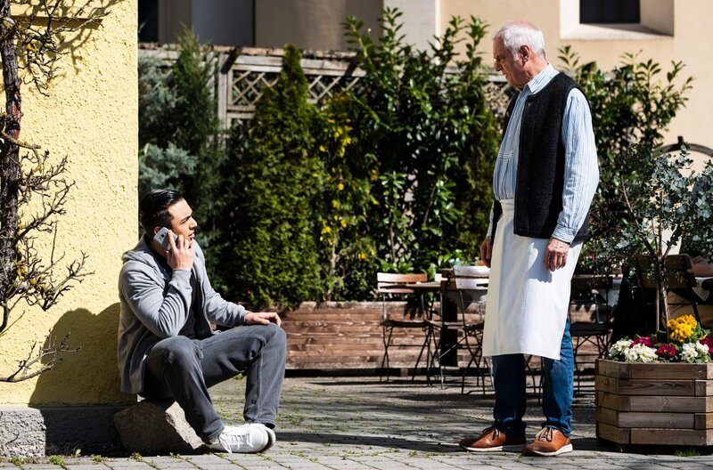 Leon (Joel Akgün) sitzt telefonierend vor dem Brunnerwirt, als Joseph (Michael Vogtmann) auf ihn zukommt. – Bild: BR/​Lena Semmelroggen