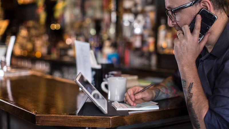 portrait of pub/​bar owner working on smartphone and electronic tablet at bar drinking coffee – Bild: Steve Prezant /​ Getty Images/​Image Source