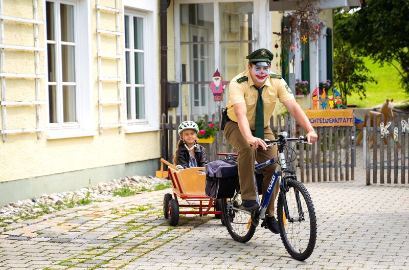 Riedl (Paul Sedlmeir, r.) bemerkt nicht, dass er aussieht wie ein Clown und macht sich mit Lina (Carla Graßl, l.) im Bollerwagen auf den Weg zum Revier. – Bild: ARD/​Jenrick Mielke