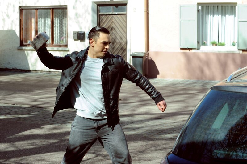 Leon (Joel Akgün) bricht in Rage ein Auto auf, das auf dem Dorfplatz steht. – Bild: Marco Orlando Pichler /​ BR /​ BR/​Marco Orlando Pichler