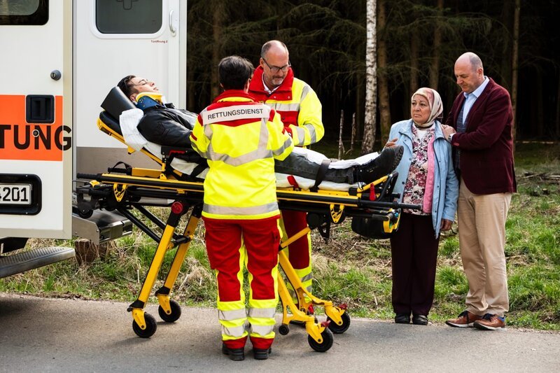Leon wird von den Rettungskräften in den Krankenwagen geladen. Von links: Leon (Joel Akgün), Rettungskräfte (Komparsen), Sevim (Özay Aslanhan) und Hubert (Bernhard Ulrich). – Bild: Lena Semmelroggen /​ BR
