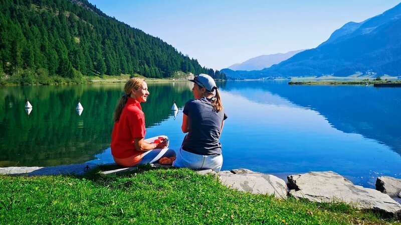 Morgens ist der Silvaplanersee noch spiegelglatt. Aber ab mittags weht hier der Malojawind, erfährt Tamina Kallert von der Surflehrerin Sarah Missiaen (r). – Bild: WDR/​Anja Koenzen