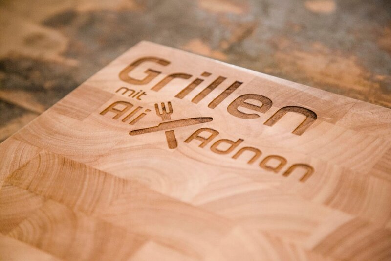 "Grillen mit Ali und Adnan"-Sendungslogo auf einem Holzbrett. – Bild: BR "Grillen mit Ali und Adnan"-Sendungslogo auf einem Holzbrett. – Bild: BR