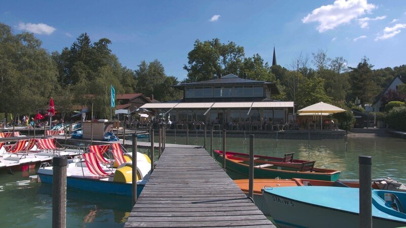 Blick auf das Seehaus Raabe am Wörthsee. Im Vordergrund stehen bunte Boote an einem Bootssteg. – Bild: BR/​Bewegte Zeiten Filmproduktion GmbH/​Alica Reisner