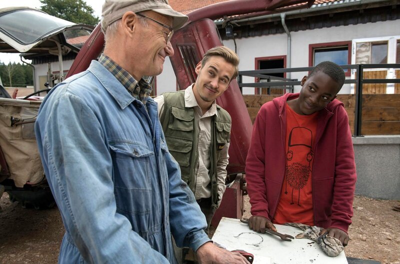 Tayo (Charles Henri Junior, re.) ist inzwischen jedem in der Familie sehr ans Herz gewachsen. Karl (Peter Schell, li.) repariert mit ihm den Anlasser vom Schlepper, was Sebastian (Dominik Stricker, Mi.) auch interessiert. – Bild: SWR/​Alexander Kluge