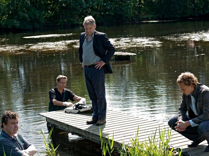 Kommissar Wallander (Kenneth Branagh, 2. v. re.) und seine Kollegen Martinsson (Tom Hiddelston, re.) und Nyberg (Richard McCabe, li.) untersuchen einen Tatort, an dem ein Mann auf grausame Weise ertränkt wurde. – Bild: NDR/​Degeto/​Yellow Bird