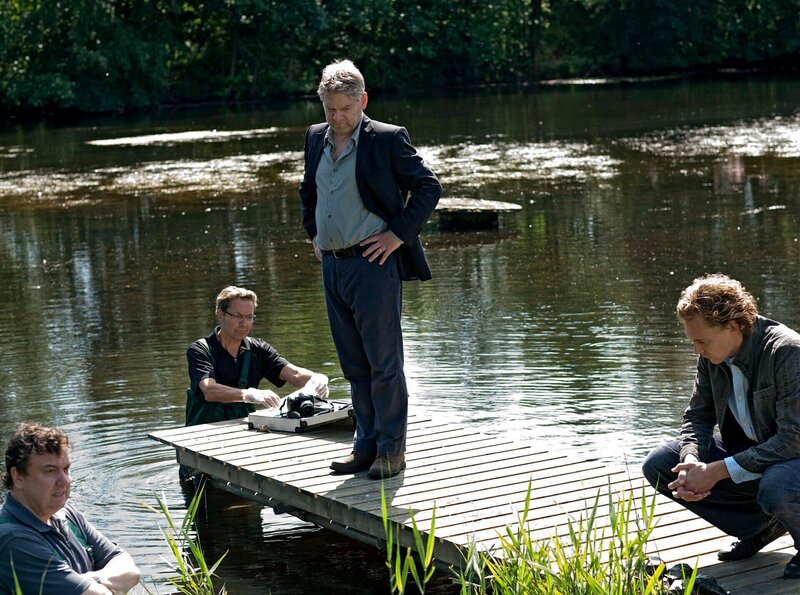 Kommissar Wallander (Kenneth Branagh, 2. v. re.) und seine Kollegen Martinsson (Tom Hiddelston, re.) und Nyberg (Richard McCabe, li.) untersuchen einen Tatort, an dem ein Mann auf grausame Weise ertränkt wurde. – Bild: NDR/​Degeto/​Yellow Bird