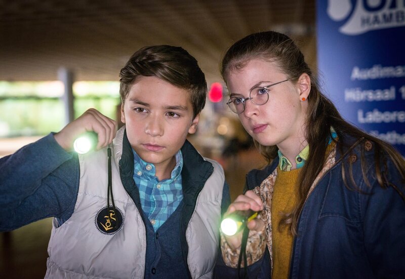 Ramin (Jann Piet, li.) und Pinja (Sina Michel, re.) beobachten das Pförtnerhaus der Hamburger Uni. – Bild: NDR/​Boris Laewen