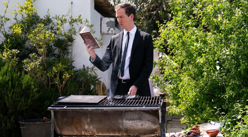 Leander Lost (Jan Krauter) greift zur besseren Verständigung auf das „Kompendium der sinnlosen Sätze“ zurück. – Bild: ARD Degeto/​Mariella Koch /​ ARD Degeto/​Programmplanung und P