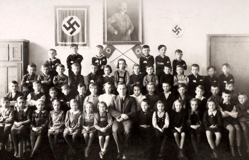 Ein Klassenporträt von jungen Schulkindern mit ihrem Lehrer vor einer Fotografie von Adolf Hitler und zwei Hakenkreuzen während des Zweiten Weltkriegs, um 1940. – Bild: TVNOW