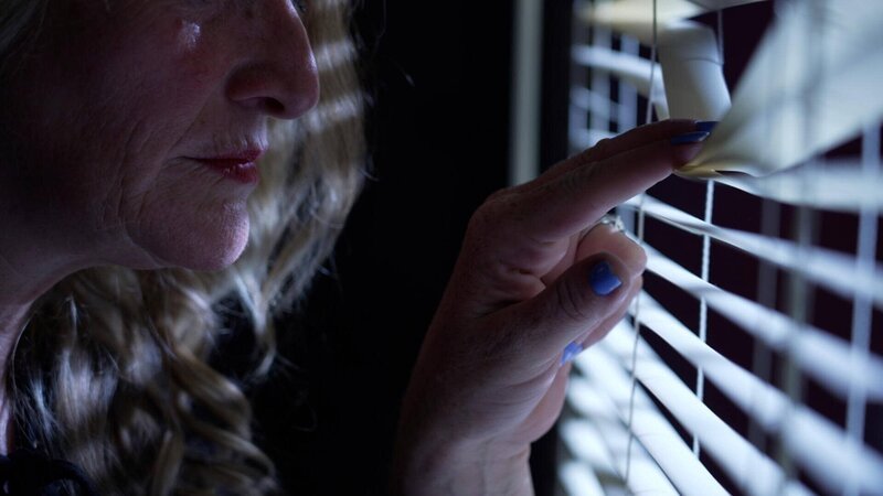Eine Frau schaut durch Jalousien. – Bild: Crime + Investigation /​ fairpoint