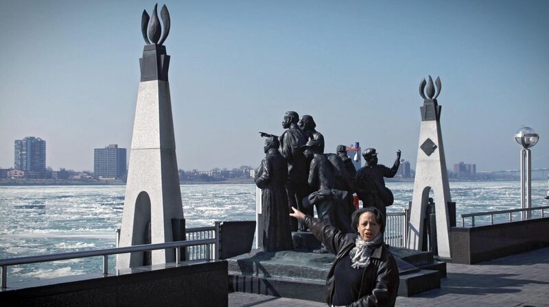 Kimberly Simmons am Denkmal der „Underground Railroad“ zeigt auf die andere Uferseite. Dort hin waren auch einst ihre Vorfahren vor der Sklaverei geflüchtet, über den Detroit River, ins freie Kanada. – Bild: 3sat