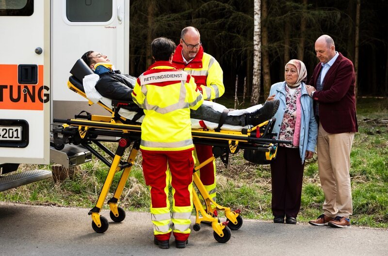 Leon wird von den Rettungskräften in den Krankenwagen geladen. Von links: Leon (Joel Akgün), Rettungskräfte (Komparsen), Sevim (Özay Aslanhan) und Hubert (Bernhard Ulrich). – Bild: BR/​Lena Semmelroggen