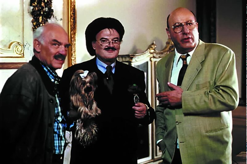 Stoever (Manfred Krug, rechts) und Brockmöller (Charles Brauer, links) ziehen Erkundigungen über die Familie der Schönach und Rataus ein. Der schillernde Titelhändler Bechtel (Rudolph Moshammer) hilft ihnen weiter. Hund Daisy zeigt sich teilnahmslos. – Bild: ORF (FS1/​FS2)