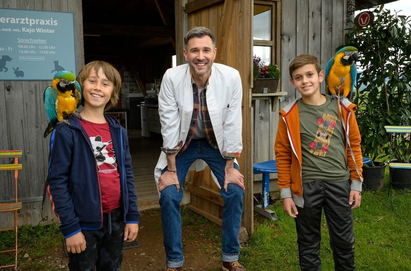 Sam (Yann Cosack, li.), Liam (Nils Hofschneider, re.) und Papa Kajo (Jochen Schropp, mi.) vor der Tierarzt-Praxis mit zwei Papageien – Bild: ARD/​Maria Wiesler