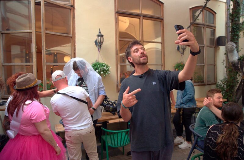 Reporter Frederik Fleig filmt sich bei einer Moderation in einer Kneipe in Prag. Im Hintergrund stehen Junggesellengruppen. – Bild: MDR/​Michal Novotný