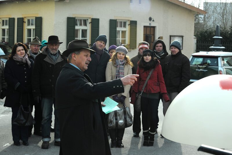 Bürgermeister Schattenhofer (Werner Rom) enthüllt das Kunstwerk, das künftig den Dorfplatz schmücken soll. – Bild: BR/​Marco Orlando Pichler