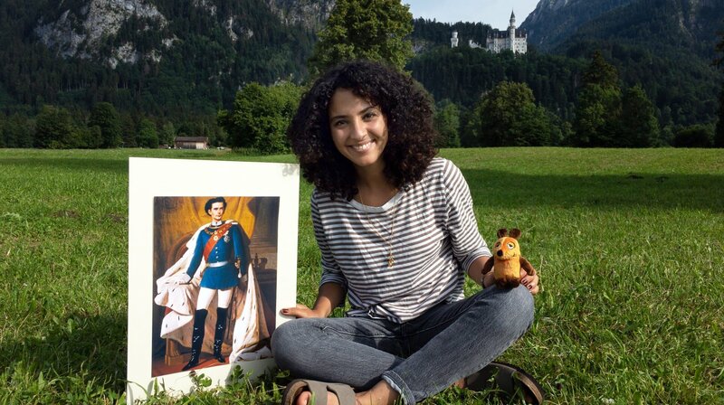 Moderatorin Siham El Maimouni mit einem Bild von König Ludwig vor dem Schloss Neuschwanstein – Bild: WDR/​gomie