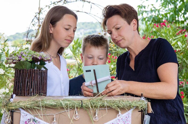 Petra Rapp (re.) packt mit ihren Kindern Leonie (li.) und Jonas eine Überraschungskiste aus. – Bild: SWR/​megaherz/​Marketa Novotna