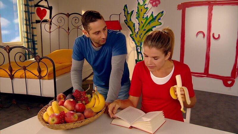 Denis Moschitto würde sich so gerne eine leckere Banane von Anke Engelkes Obstteller nehmen, doch dafür muss er zuerst das Zauberwort nennen. – Bild: WDR/​Trickstudio Lutterbeck