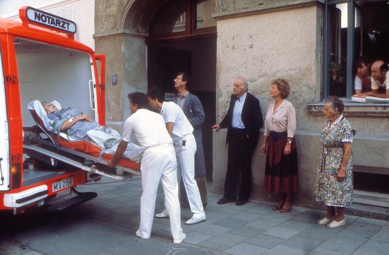 Die Hausbesitzerin Frau Winter (Erika Wackernagel, links) muß ins Krankenhaus. Von links: Ihr Neffe, Herr Eggerer (Gerhard Zemann), der Mieter Herr Schrambeck (Helmut Brasch), die Hausmeisterin Martha Haslbeck (Veronika Fitz), eine Anwohnerin (Komparsin) sowie die Mieter Rosa (Sarah Camp) und Hans Ostermeier (Hans Stadlbauer) beobachten das traurige Ereignis. – Bild: Bayerischer Rundfunk