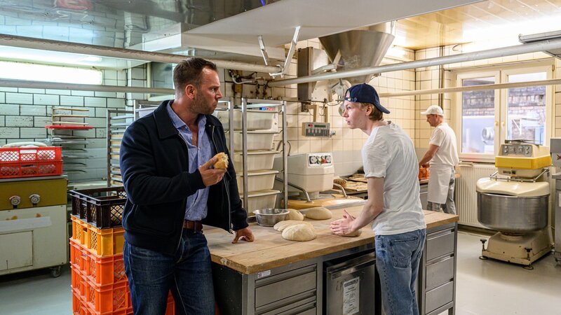 Luis Braun (Filip Schnack, M.) führt Kommissar Lars Pöhlmann (Dominic Boeer, l.) seine Backkünste vor. Mit seinen Anteilen an der Firma will er die Bäckerei auf bio umstellen. Doch seine Tante, die ebenfalls am Unternehmen beteiligt ist, sieht darin eine Gefahr: Für sich und die Firma. Es stellt sich die Frage: Wer hat das Sagen in der Großbäckerei? Dabei könnten beide von Peter Brauns Tod profitieren. – Bild: ZDF