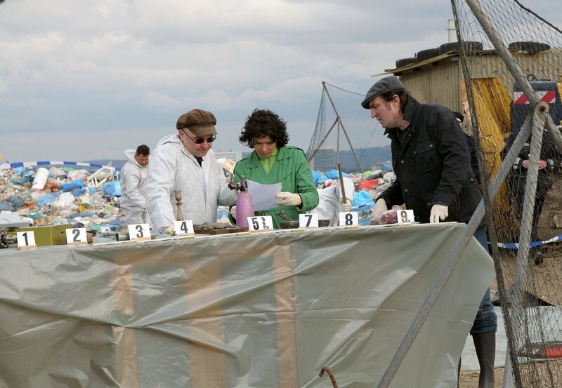 Peter Becker hat am übelriechenden Fundort der Leiche, einer Müllkippe, eine Sammlung von denkbaren Tatwaffen für Lena Odenthal (Ulrike Folkerts) und Kopper (Andreas Hoppe) aufgebaut. – Bild: SWR/​Krause