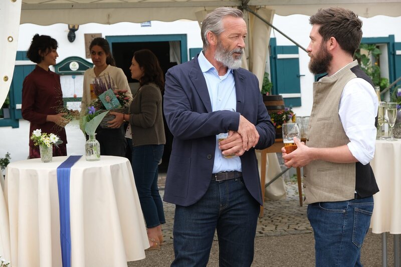 Vinz Huber (Michael Roll, M.) und Schorschi Striebel (Sebastian Edtbauer, r.) haben sehr unterschiedliche Auffassungen darüber, was den Umgang mit der stillgelegten Haltestelle in Himmelsruh betrifft. Im Hintergrund stehen Charlie Tanaka (Agnes Decker, l.), Julia Obermeier (Liane Forestieri, 2.v.l.) und Hanne Striebel (Kerstin Dietrich, 3.v.l.) zusammen. Hanne hält einen Geschenkkorb in der Hand. – Bild: ZDF