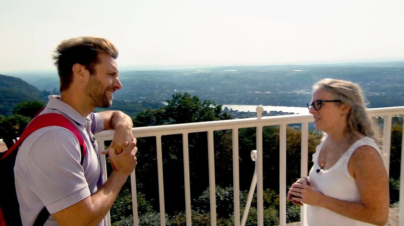 Soledad Sichert (r), Simultandolmetscher und Reiseführerin, erzählt Daniel Aßmann auf dem Balkon des Luxushotels Petersberg Anekdoten über die prominenten Gäste des ehemaligen Gästehauses des Bundes. – Bild: BR/​WDR