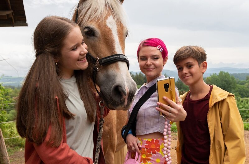 Lucy (Paulina Schnurrer, links) und Liam (Nils Hofschneider, rechts) sind superglücklich. Ihre Lieblings-Influencerin Tara Lovely (Carlotta Truman, Mitte) ist zu Besuch in Waldau. – Bild: SWR/​ARD/​Maria Wiesler