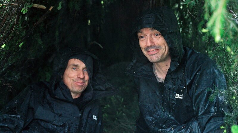 v.li.: Marco und Armin versuchen sich vor dem Regen zu schützen. Die gute Laune vergeht ihnen aber bald. Langsam wird es kalt und bis jetzt haben sie noch kein Feuer hingekriegt. – Bild: SRF