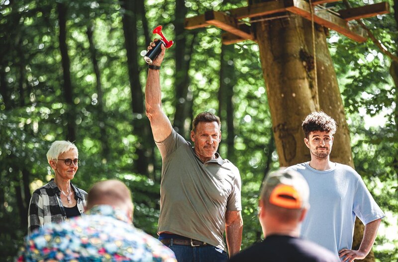 Ralf Moeller startet mit dem Luftdruckhorn die Uhr bei Motorsägen Masters. Sylvia Itzen und Tim Bengel sind gespannt auf die Kunstwerke der Teilnehmenden. – Bild: SWR