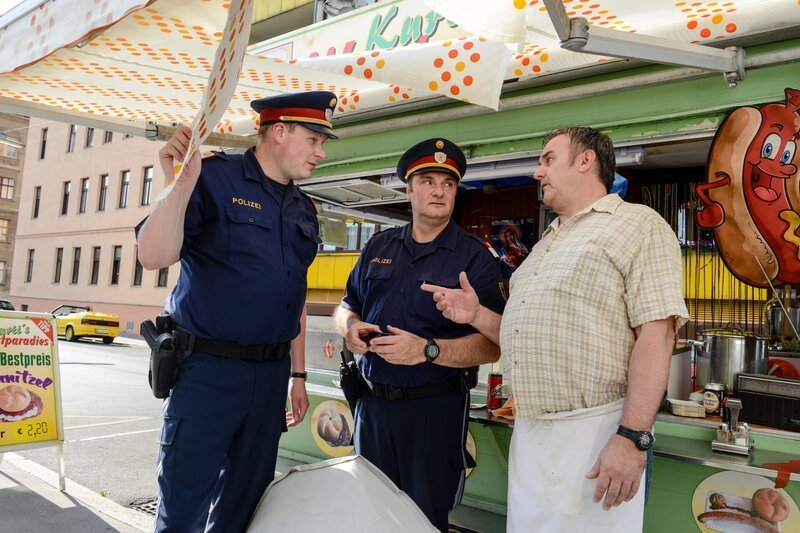„CopStories“, „Haaße Luft.“ Sommerliche Hitze hat die Stadt fest im Griff. Da hat die Polizei mehr als sonst zu tun: Eine Mutter bringt zur Anzeige, dass ein unbekannter Mann ihrer Tochter im Kindergarten Haare abgeschnitten hat, ein Würstelstand wird von herabgeworfenen Gegenständen demoliert und in einem „Massagesalon“ kommt es zum wahrscheinlich seltsamsten Fall von Samenraub. Währenddessen gehen die Ermittlungen im Fall der ermordeten Swetlana auf Hochtouren weiter. Erschwert werden sie dadurch, dass der Verdacht aufkommt, dass ein korrupter Polizist in den Fall verwickelt sein könnte.Im Bild (v.li.): Holger Schober (Roman Mohácsi), Martin Leutgeb (Mathias Gerber), Christian Dungl (Würstelstandler). SENDUNG: ORF eins – DI – 02.10.2018⁠–⁠21:05 UHR. – Bild: ORF/​Hubert Mican