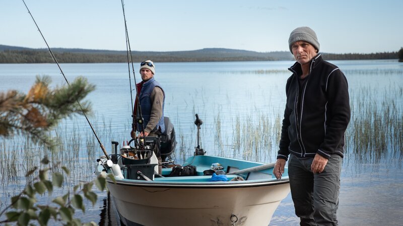 Jeremy Wade standing next to a moored boat on the edge of a lake, Guide Oscar Hedskog standing on the back of the boat next to the engine. Foliage out of focus in foreground. – Bild: Discovery Communications, LLC