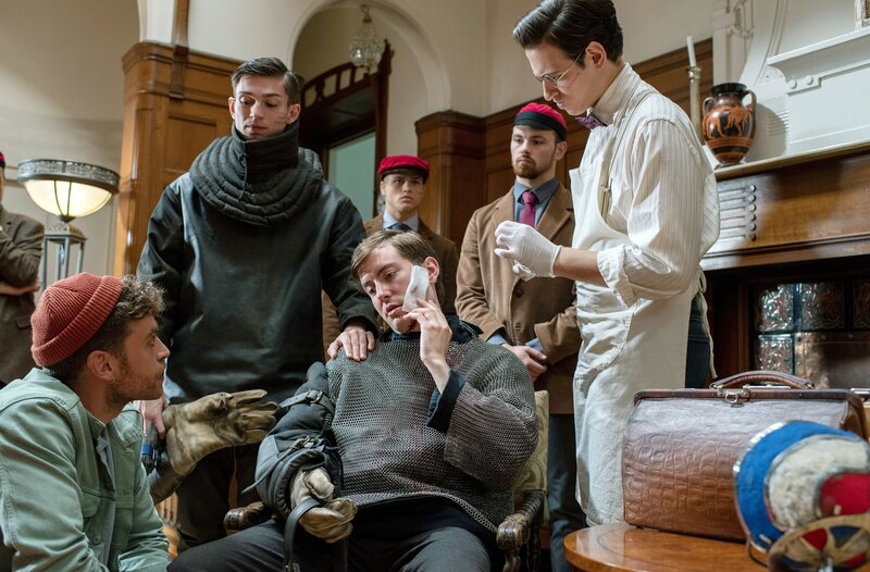 Beim Fechtkampf in der Burschenhaft wurde August Schöll (Lukas Nathrath, M.) schwer am Kopf und im Gesicht verletzt. Auf Nils Sanchez (Enrique Fiß, l.) wirkt der verletzte Student benebelt, doch Marko Bremer (Vincent zur Linden, 2.v.l.) wiegelt Hilfe und Nachfragen ab (mit Komparsen). – Bild: ARD/​Thorsten Jander